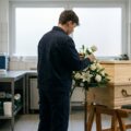 Auszubildende richtet in einem Bestattungsinstitut Blumen neben einem einfachen Holzsarg.