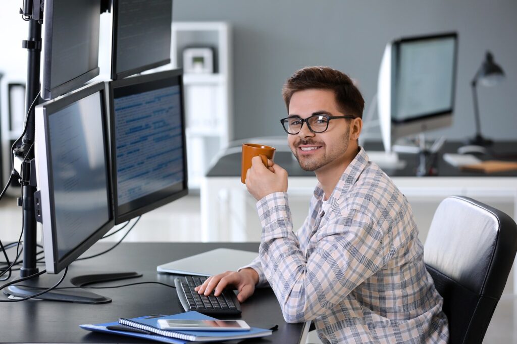 Junger Softwareentwickler mit Kaffeetasse an einem Arbeitsplatz mit mehreren Monitoren voller Code