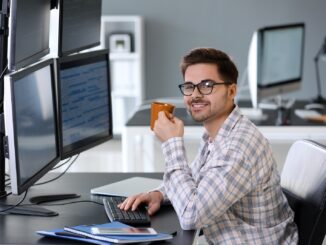 Junger Softwareentwickler mit Kaffeetasse an einem Arbeitsplatz mit mehreren Monitoren voller Code