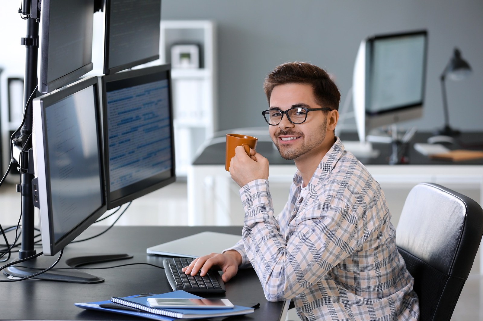 Junger Softwareentwickler mit Kaffeetasse an einem Arbeitsplatz mit mehreren Monitoren voller Code